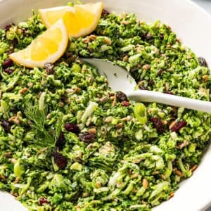 A spoon resting in a bowl of broccoli slaw garnished with lemon wedges.