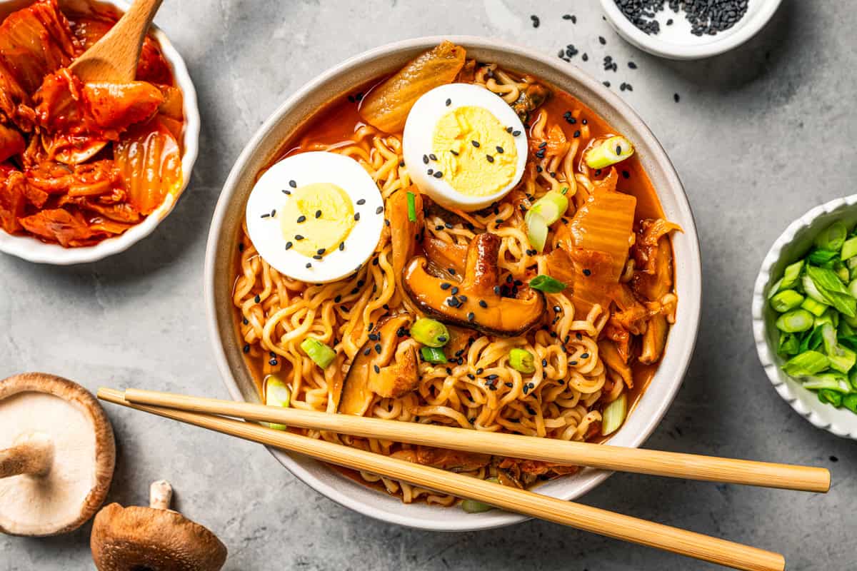 This quick and easy kimchi ramen recipe packs a spicy, savory punch with instant noodles, kimchi, shiitake mushrooms, and gochujang broth. A bowl of spicy kimchi ramen with egg, mushrooms, and green onions, with chopsticks on the side.
