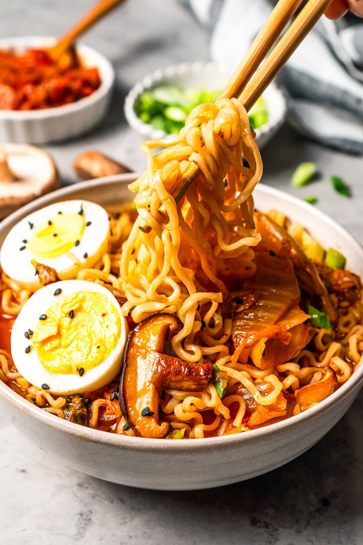 This quick and easy kimchi ramen recipe packs a spicy, savory punch with instant noodles, kimchi, shiitake mushrooms, and gochujang broth. Close up of chopsticks lifting noodles from a bowl of Korean kimchi ramen with egg.