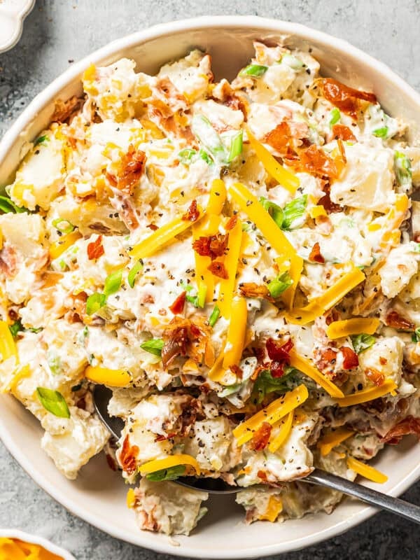 A spoon resting in a bowl of loaded baked potato salad with bacon and cheddar.
