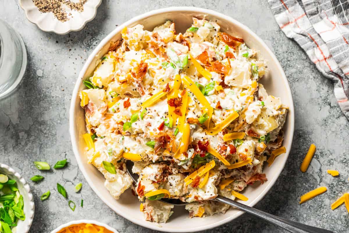 This rich and creamy loaded potato salad with bacon, spring onions, and cheddar cheese is easy to make ahead, perfect for potlucks and BBQs! A spoon resting in a bowl of loaded baked potato salad with bacon and cheddar.