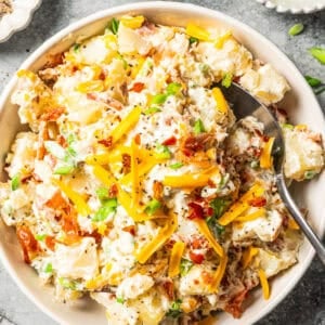A spoon resting in a bowl of loaded baked potato salad with bacon and cheddar.