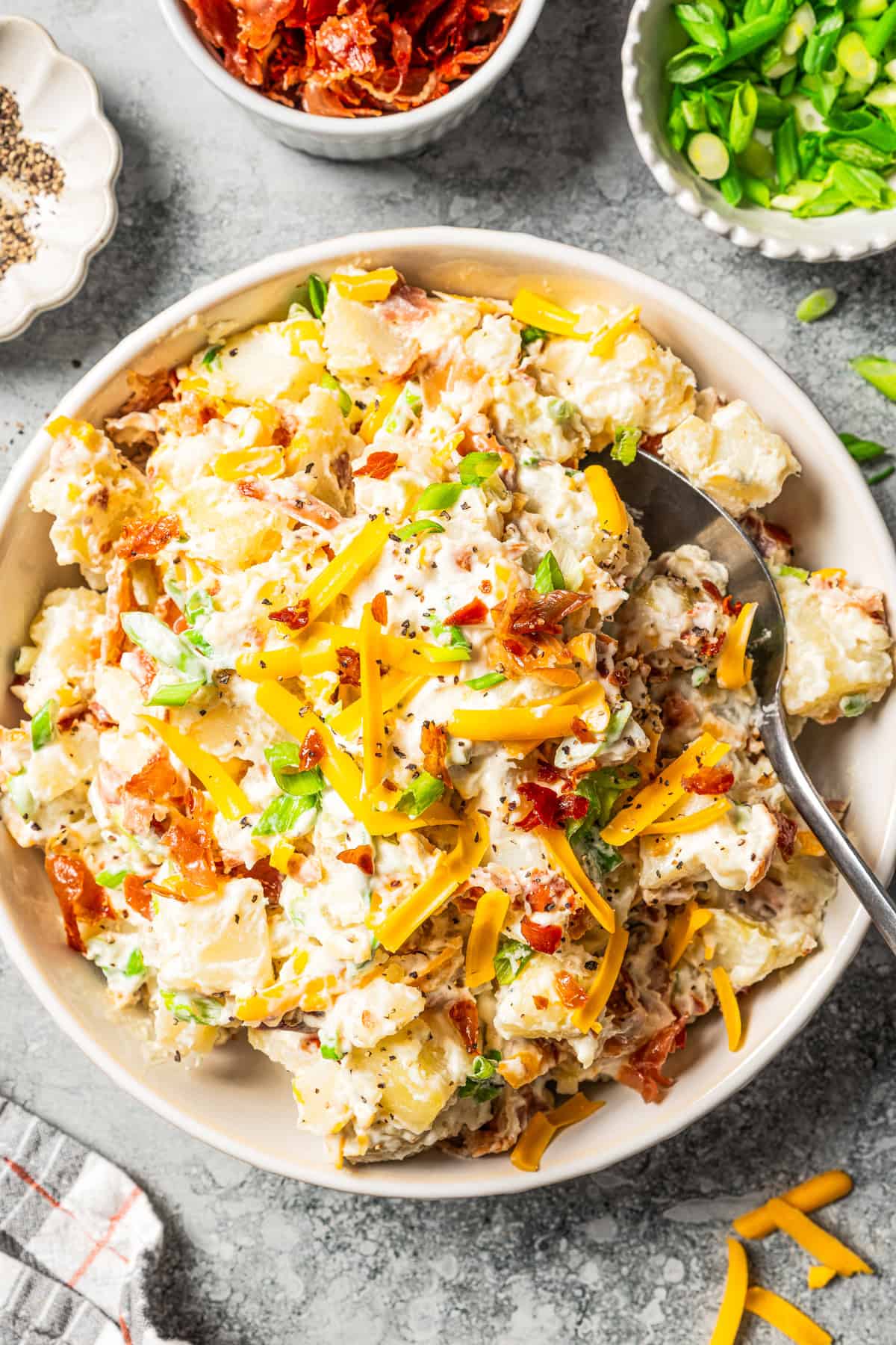 A spoon resting in a bowl of loaded baked potato salad with bacon and cheddar.