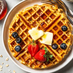 A protein waffle topped with fresh fruit, butter, and maple syrup on a plate.