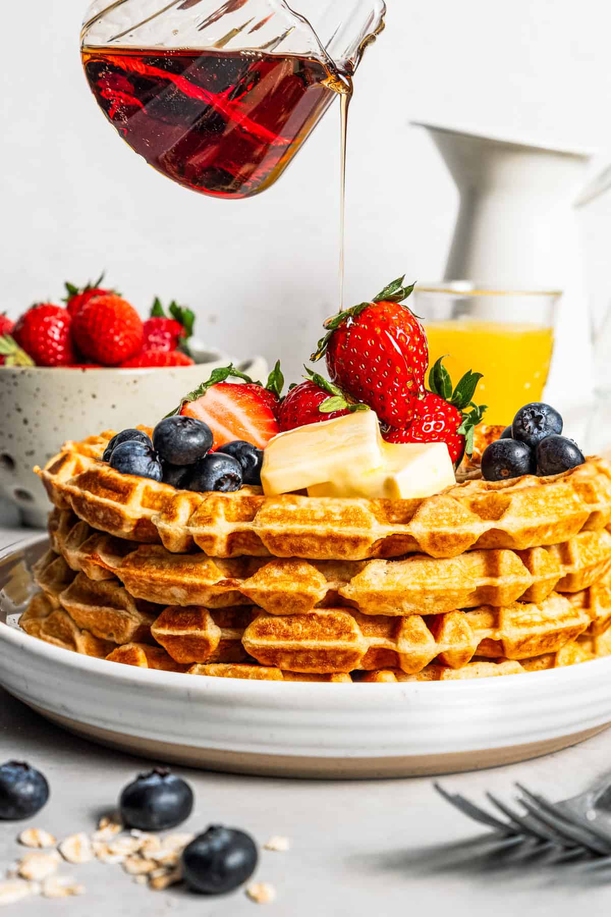 Pouring maple syrup over a stack of protein waffles topped with berries and butter on a plate.
