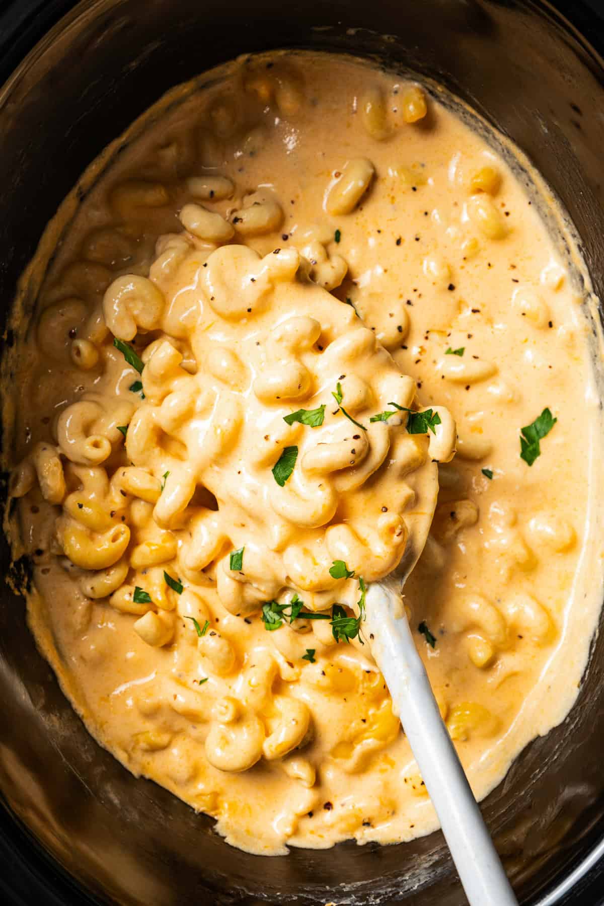 Ultra-creamy slow cooker mac and cheese is a dump-and-go version of my homemade mac and cheese recipe! Perfect for potlucks and holidays. A spoon lifting mac and cheese from the slow cooker.