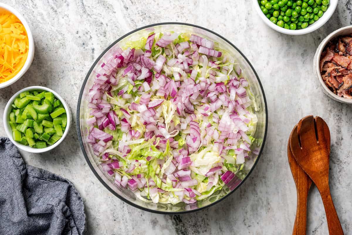 7-layer salad is a staple side dish at potlucks and BBQs! Made with crisp lettuce, onions, peas, cheese, celery, bacon, and creamy dressing. Red onions added on top of shredded lettuce in a salad bowl.