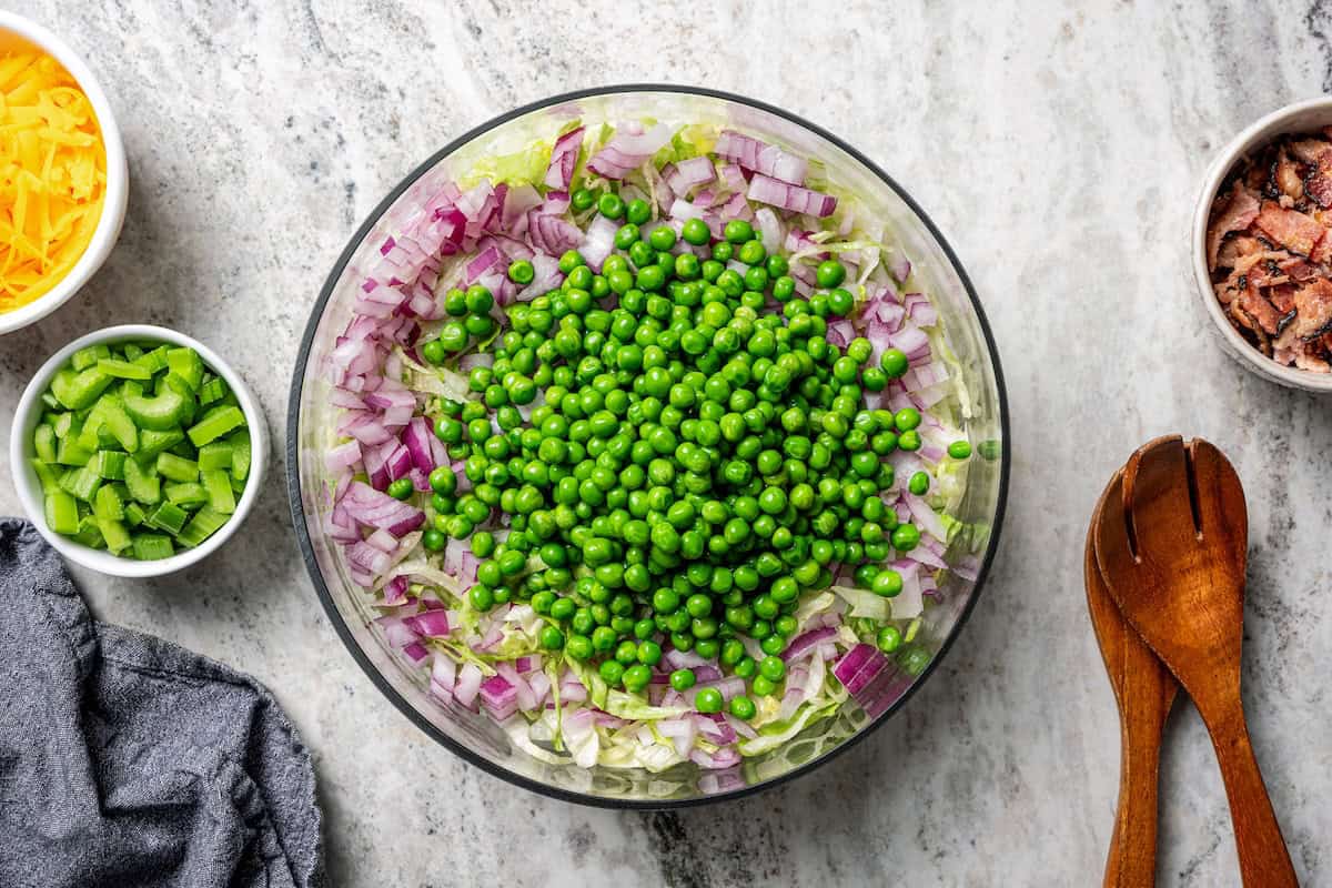 7-layer salad is a staple side dish at potlucks and BBQs! Made with crisp lettuce, onions, peas, cheese, celery, bacon, and creamy dressing. Frozen peas layered over lettuce and red onions in a salad bowl.