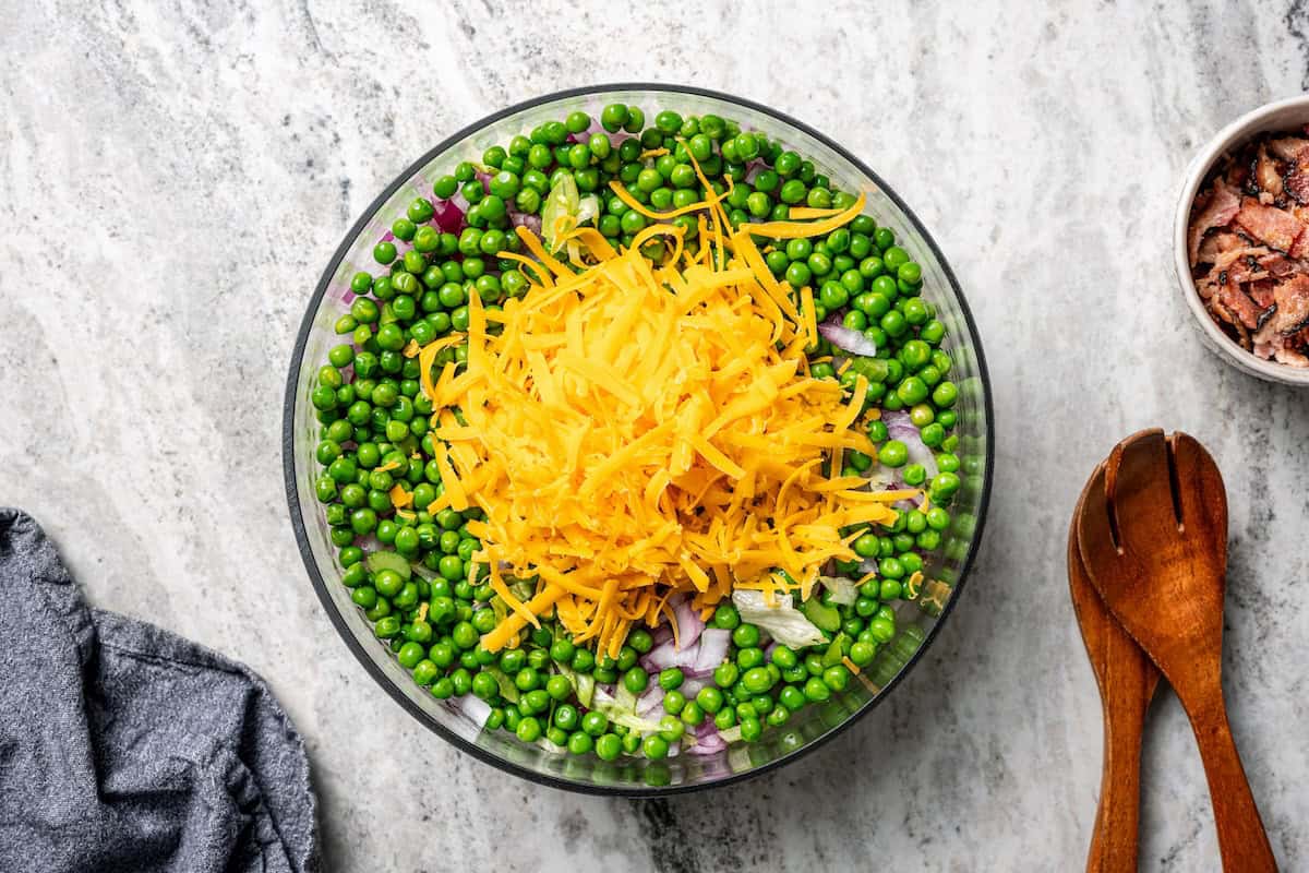7-layer salad is a staple side dish at potlucks and BBQs! Made with crisp lettuce, onions, peas, cheese, celery, bacon, and creamy dressing. Shredded cheddar cheese layered over salad ingredients in a bowl.