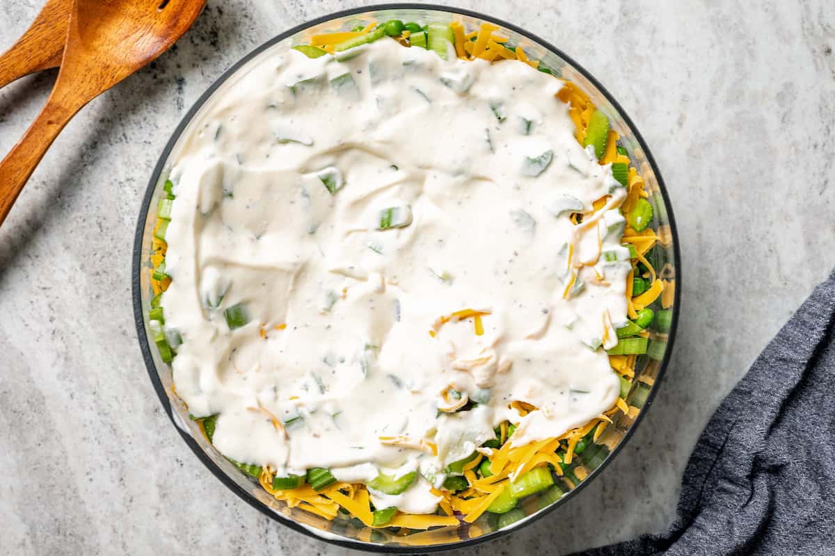 7-layer salad is a staple side dish at potlucks and BBQs! Made with crisp lettuce, onions, peas, cheese, celery, bacon, and creamy dressing. Creamy dressing spread over the salad layers in a bowl.