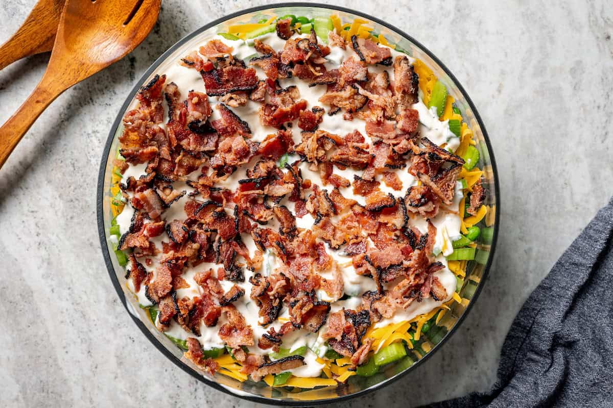 7-layer salad is a staple side dish at potlucks and BBQs! Made with crisp lettuce, onions, peas, cheese, celery, bacon, and creamy dressing. Crumbled bacon added on top of the dressing as the last layer in a 7 layer salad.