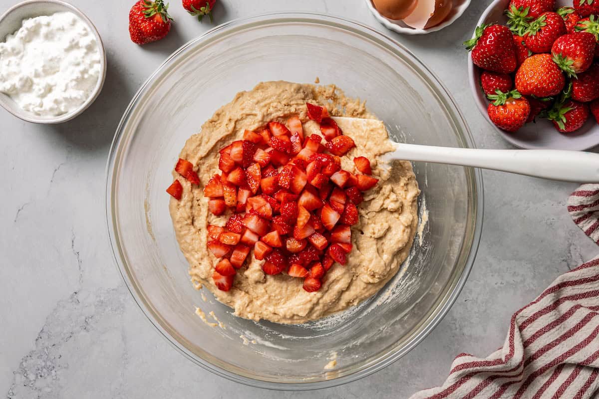 These moist strawberry protein muffins with applesauce, cottage cheese, and protein powder make an easy, light breakfast or meal prep snack! Chopped strawberries added to protein muffin batter in a bowl.