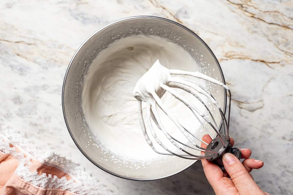 This no-bake strawberry icebox cake is an easy overnight dessert with layers of graham crackers, fresh strawberries, and fluffy whipped cream. A hand holding a whisk attachment with whipped cream over a bowl.