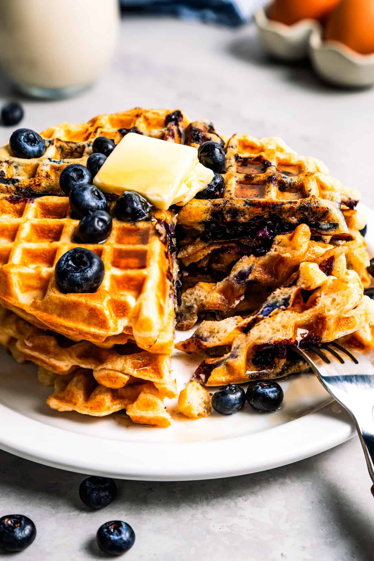 These classic blueberry waffles are light, fluffy, and ready in minutes. Rest the batter, then cook until they're golden brown and crisp. A fork cutting into a stack of blueberry waffles topped with butter on a plate.