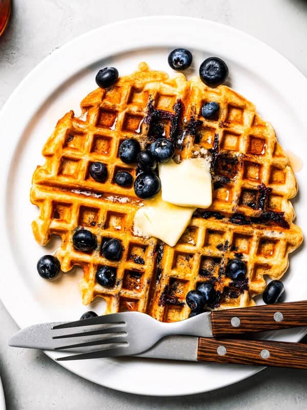 Overhead view of a fresh blueberry waffle topped with butter and maple syrup on a plate.