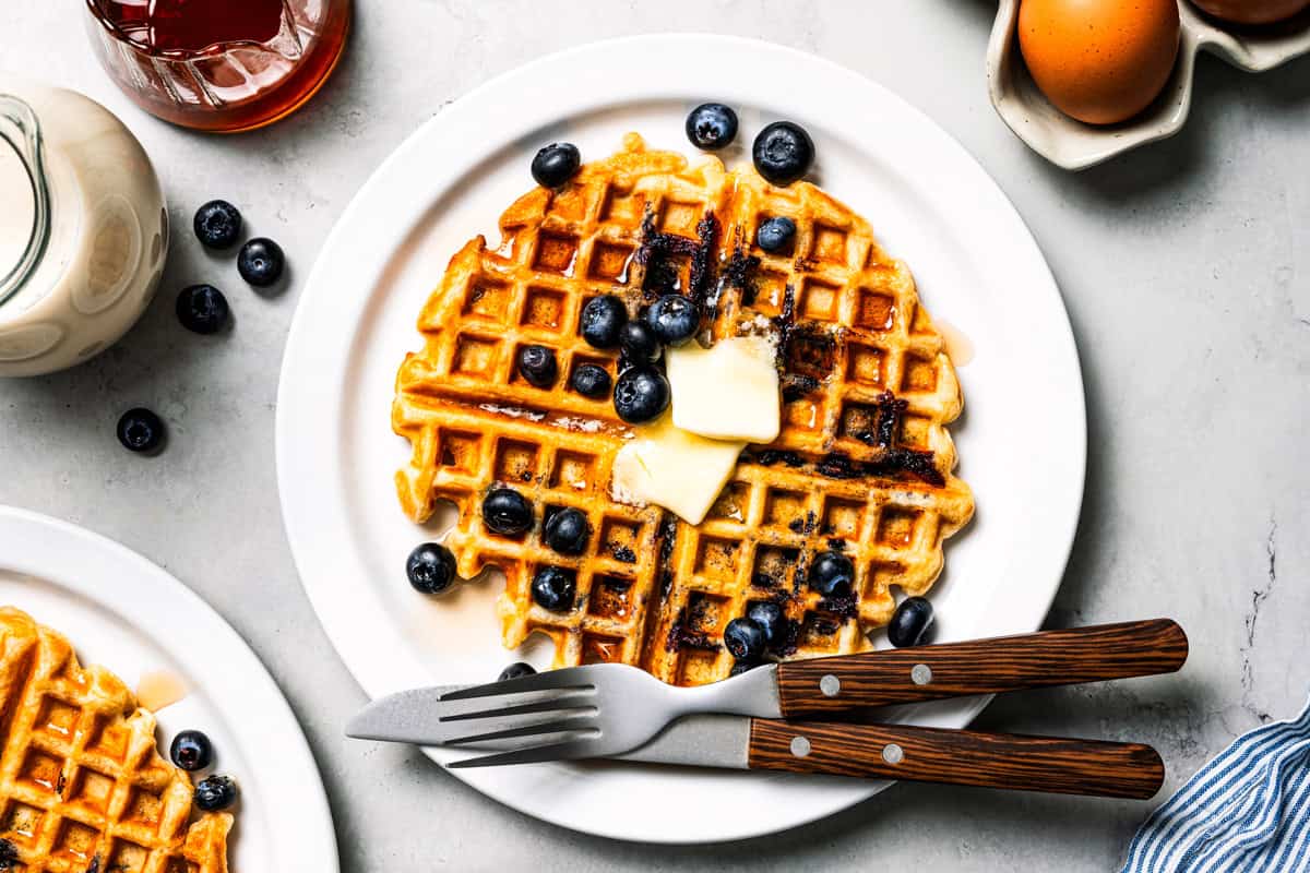 These classic blueberry waffles are light, fluffy, and ready in minutes. Rest the batter, then cook until they're golden brown and crisp. Overhead view of a fresh blueberry waffle topped with butter and maple syrup on a plate.