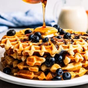 Pouring maple syrup over a stack of blueberry waffles.