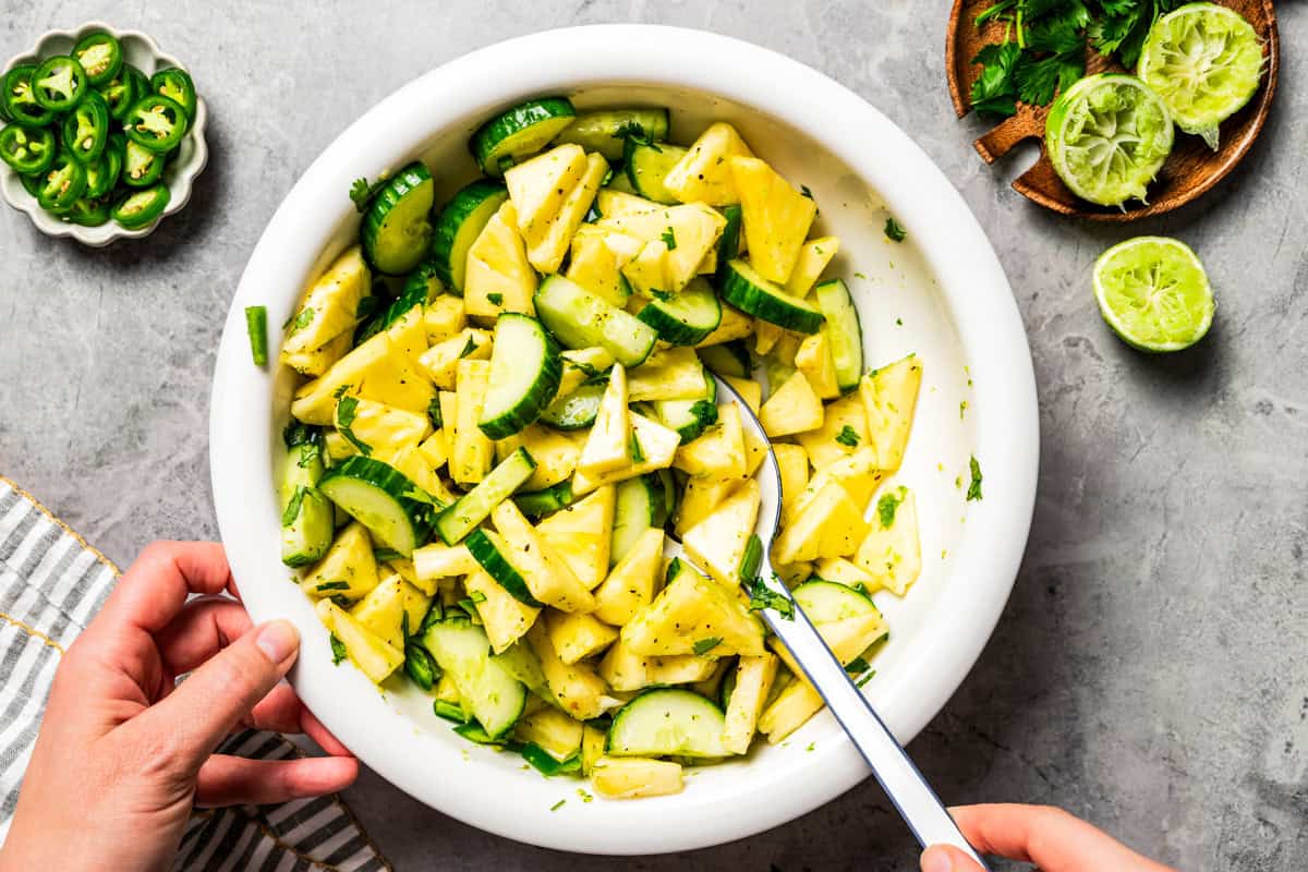 A hand stirring through a cucumber and pineapple salad in a bowl.
