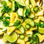 Sliced cucumbers, pineapples, lime wedges, and cilantro tossed in a salad bowl.