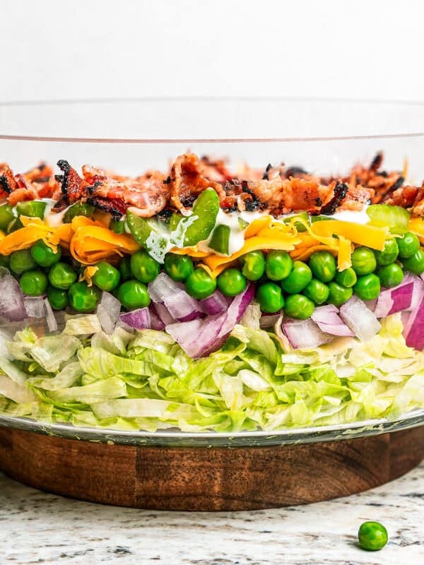 Side view of a seven layer salad with shredded lettuce, red onion, peas, shredded cheese, celery, dressing, and bacon in a glass bowl.
