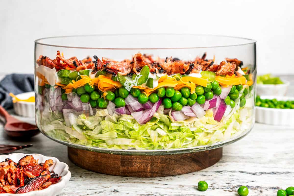 Side view of a seven layer salad with shredded lettuce, red onion, peas, shredded cheese, celery, dressing, and bacon in a glass bowl.