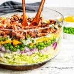 Seven layer salad with shredded lettuce, red onion, peas, shredded cheese, celery, dressing, and bacon in a glass bowl with wooden salad tongs.