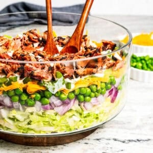 Seven layer salad with shredded lettuce, red onion, peas, shredded cheese, celery, dressing, and bacon in a glass bowl with wooden salad tongs.