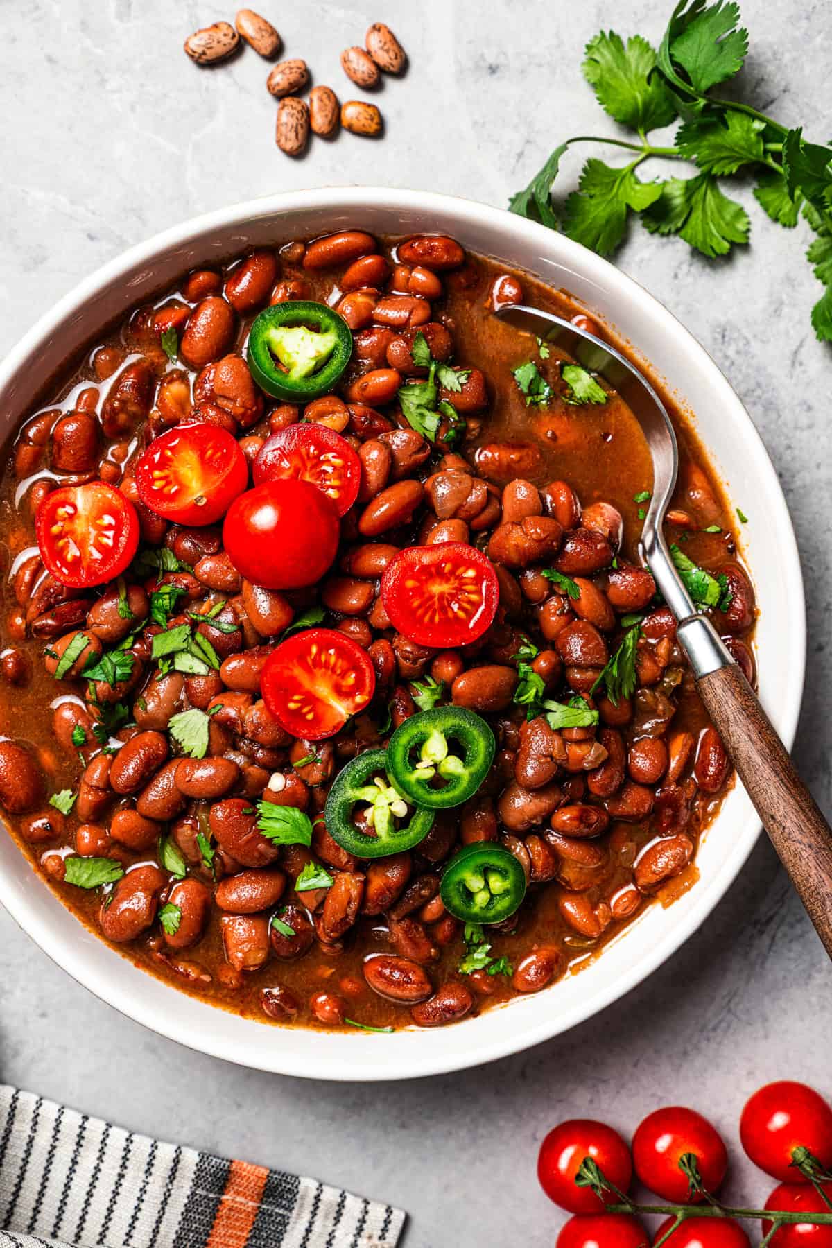 Slow cooker pinto beans are full of flavor and so easy to make. Add dry beans to the crock pot in the morning, and they’re tender by dinner! Overhead view of slow cooker pinto beans in a bowl, topped with sliced cherry tomatoes and jalapeños.