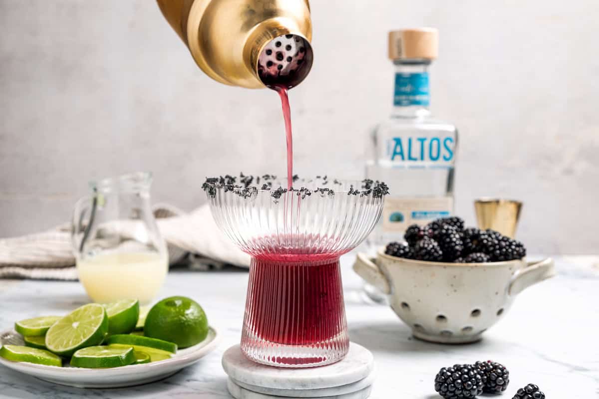 A blackberry margarita puts a refreshing, sweet-tart spin on the classic cocktail, with fresh muddled blackberries, tequila blanco, and lime. Pouring a blackberry margarita from a cocktail shaker into a salt-rimmed glass.
