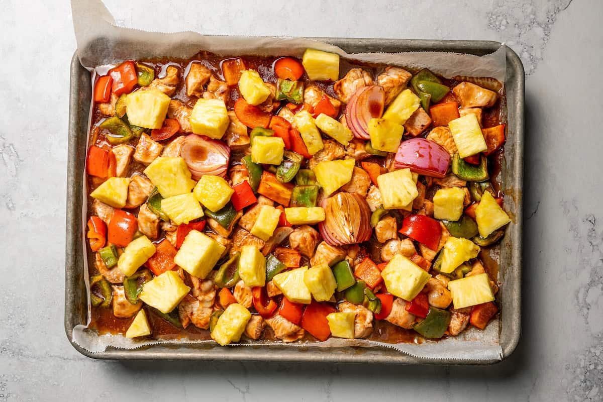 Pineapple pieces added to marinated chicken cubes and bell peppers on a baking sheet.