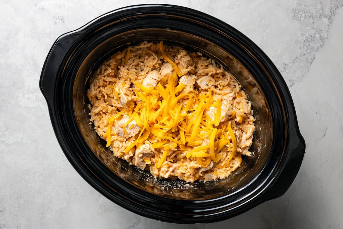 Creamy slow cooker chicken and rice is an easy one-pot dinner with rice, condensed mushroom soup, melty cheddar, onions, and juicy chicken. Shredded cheddar cheese added to creamy chicken and rice inside the slow cooker.