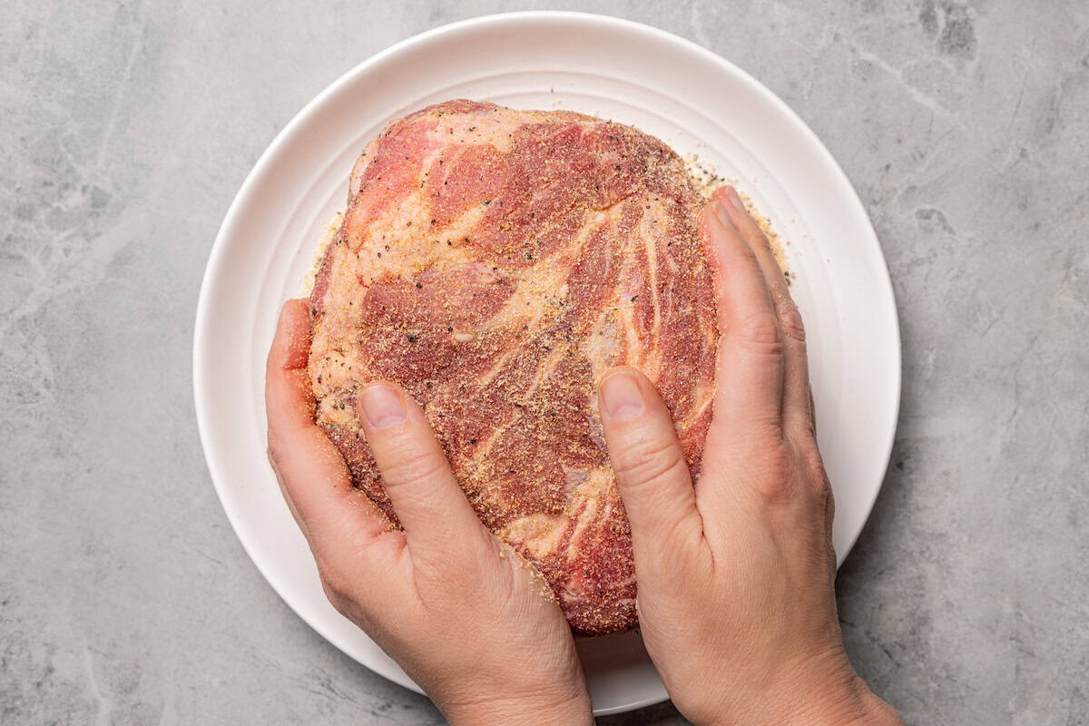 This tender, juicy slow cooker pork shoulder needs only 10 minutes of prep! It's slowly braised with hearty veggies for a cozy one-pot meal. Two hands rubbing spices over a pork shoulder.