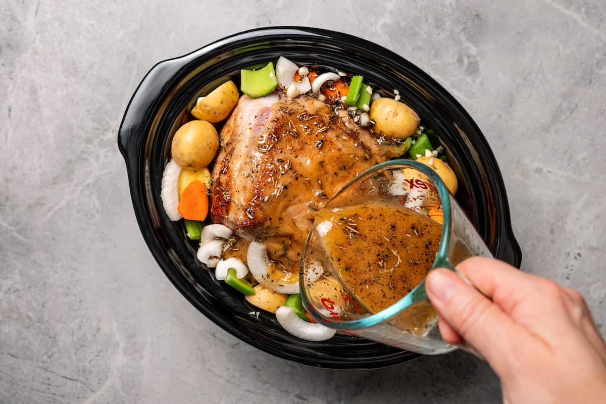 This tender, juicy slow cooker pork shoulder needs only 10 minutes of prep! It's slowly braised with hearty veggies for a cozy one-pot meal. Cooking liquid being poured over seared pork shoulder and veggies inside the slow cooker.