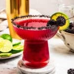 A blackberry margarita in a salt-rimmed glass garnished with fresh berries and lime slices, with a gold cocktail shaker in the background.
