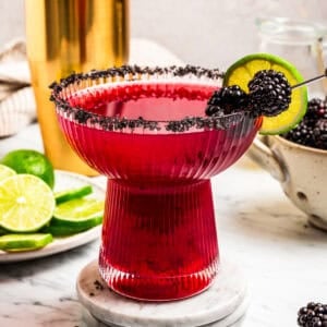 A blackberry margarita in a salt-rimmed glass garnished with fresh berries and lime slices, with a gold cocktail shaker in the background.