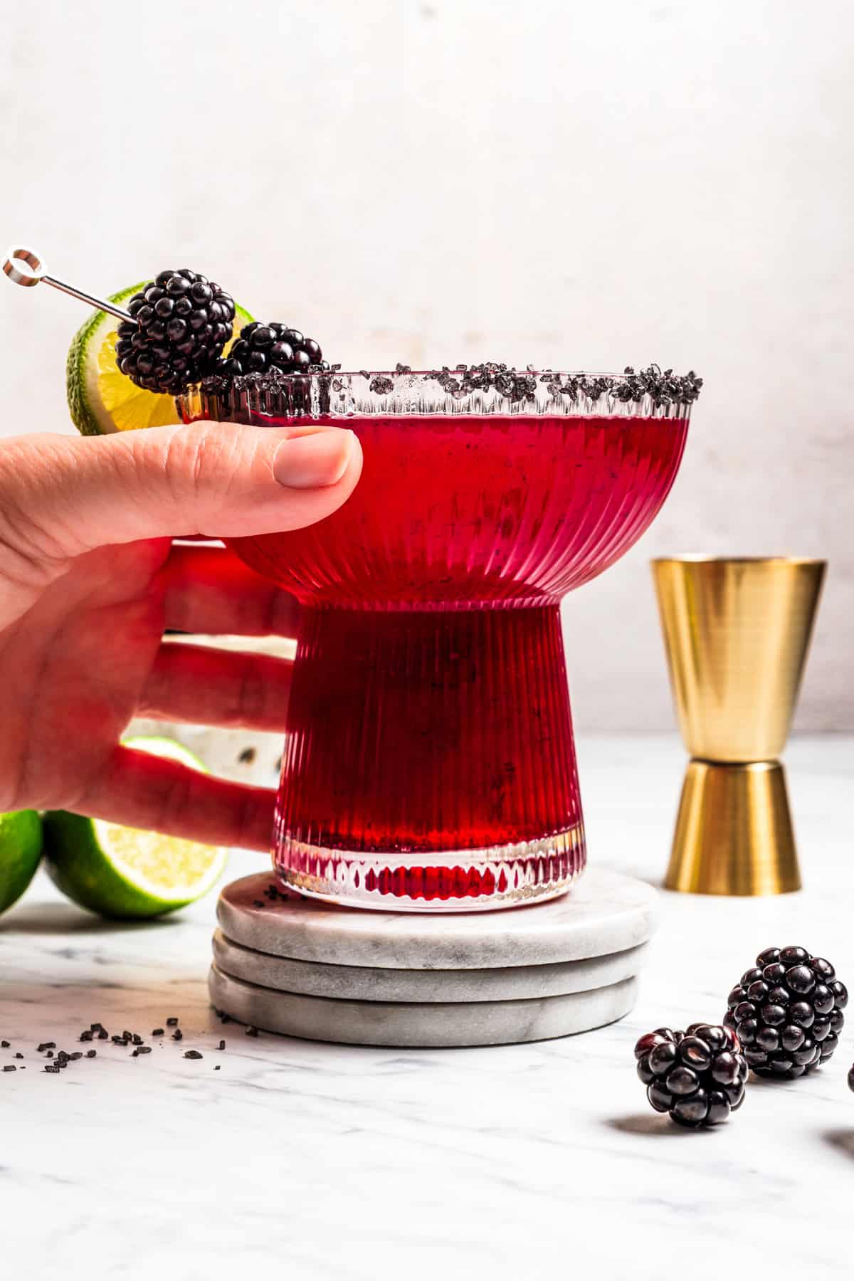 A blackberry margarita puts a refreshing, sweet-tart spin on the classic cocktail, with fresh muddled blackberries, tequila blanco, and lime. A hand reaching for a blackberry margarita on a coaster.