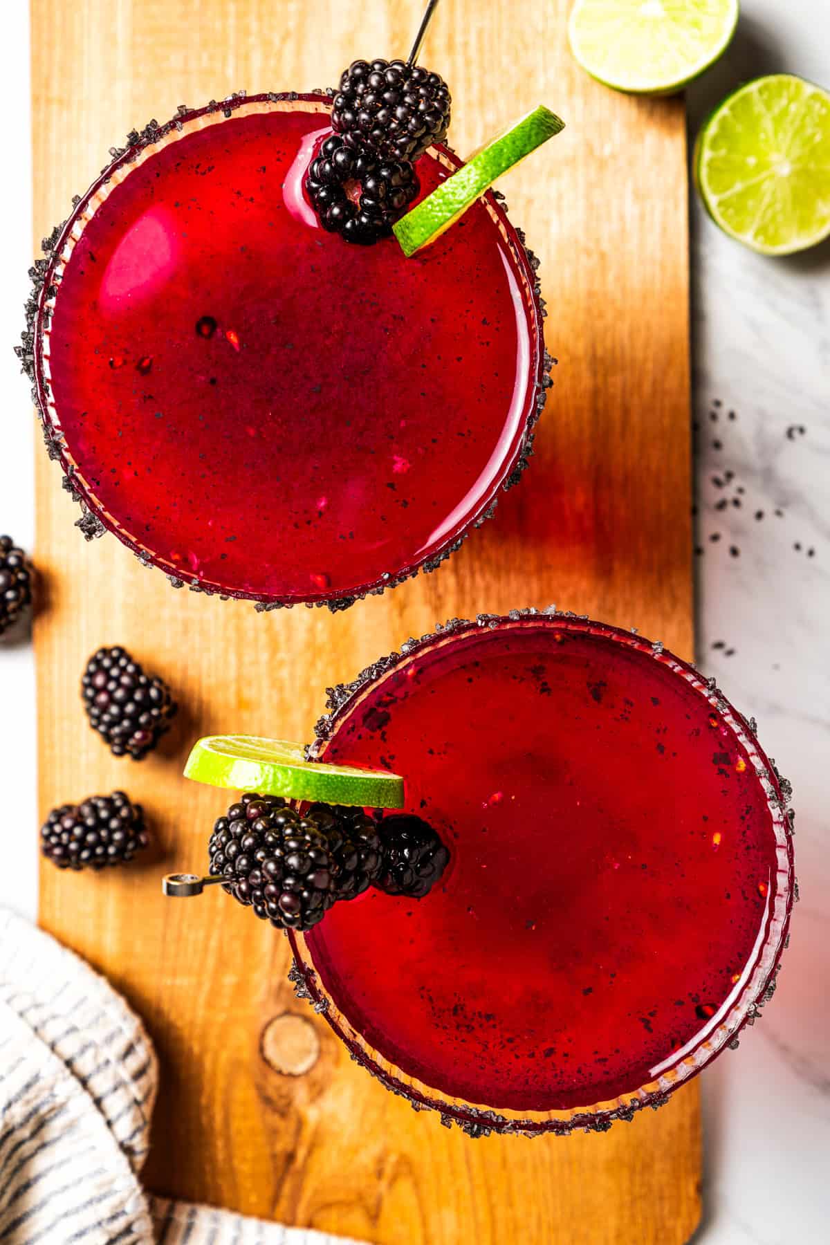 A blackberry margarita puts a refreshing, sweet-tart spin on the classic cocktail, with fresh muddled blackberries, tequila blanco, and lime. Overhead view of two blackberry margaritas in salt-rimmed glasses garnished with fresh berries and lime slices.