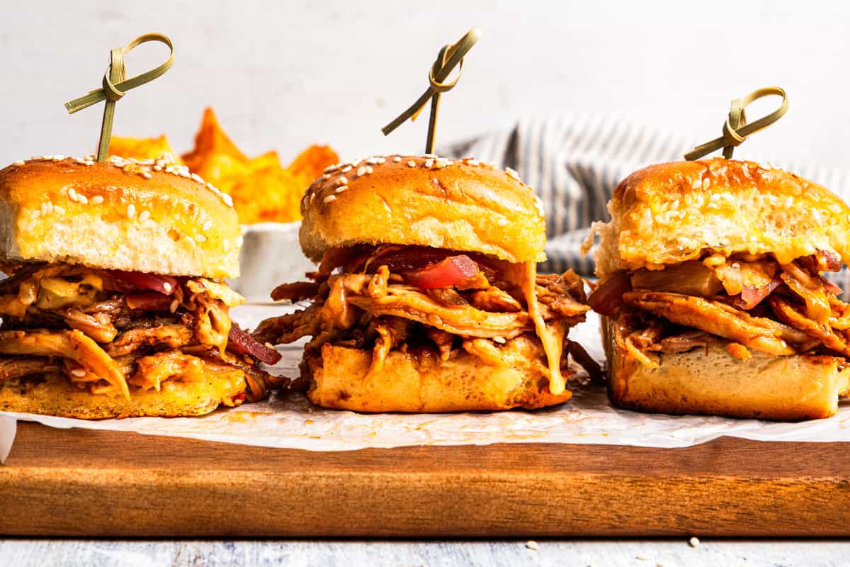 These BBQ pulled pork sliders are sweet, smoky, savory, and easy to make with leftover pulled pork, melted cheese, and fluffy Hawaiian rolls. Three pulled pork sliders on a parchment-lined wooden board.