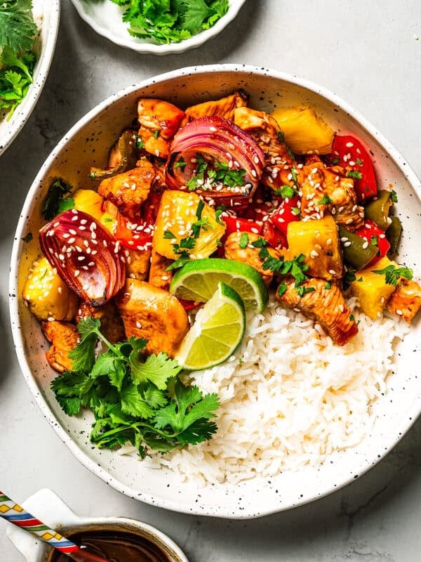 A bowl of sheet pan Hawaiian chicken featuring rice topped with chicken bites, bell pepper slices, onion slices, and pineapple chunks.