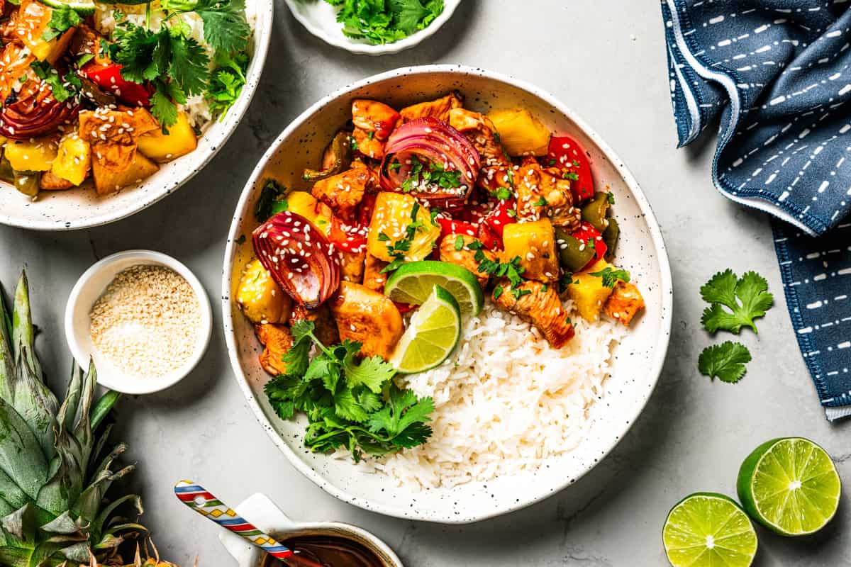 A bowl of sheet pan Hawaiian chicken featuring rice topped with chicken bites, bell pepper slices, onion slices, and pineapple chunks.