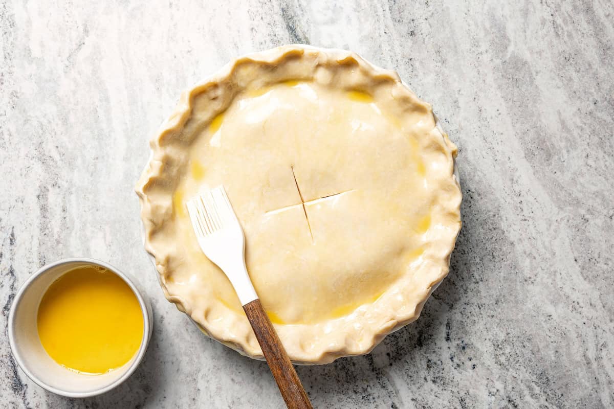 Apple berry pie is the perfect dessert to go from summer to fall! A cozy homemade apple pie bursting with fresh berries in a flaky pie crust. Brushing egg wash over the top crust of homemade apple berry pie.