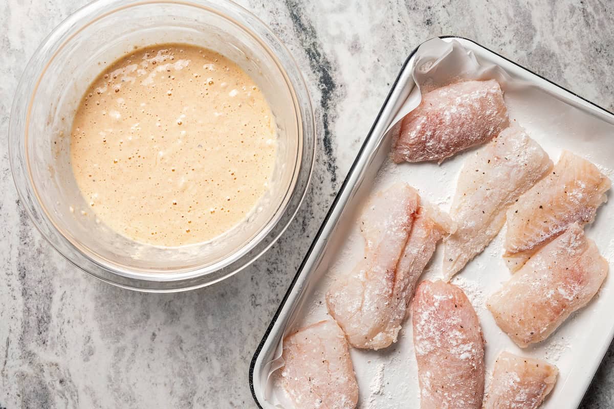 Make beer-battered fish with easy ingredients! This crispy, flaky fried white fish is perfect for tacos, pub-style fish and chips, and more. Fish filets on a sheet pan next to a bowl of batter.