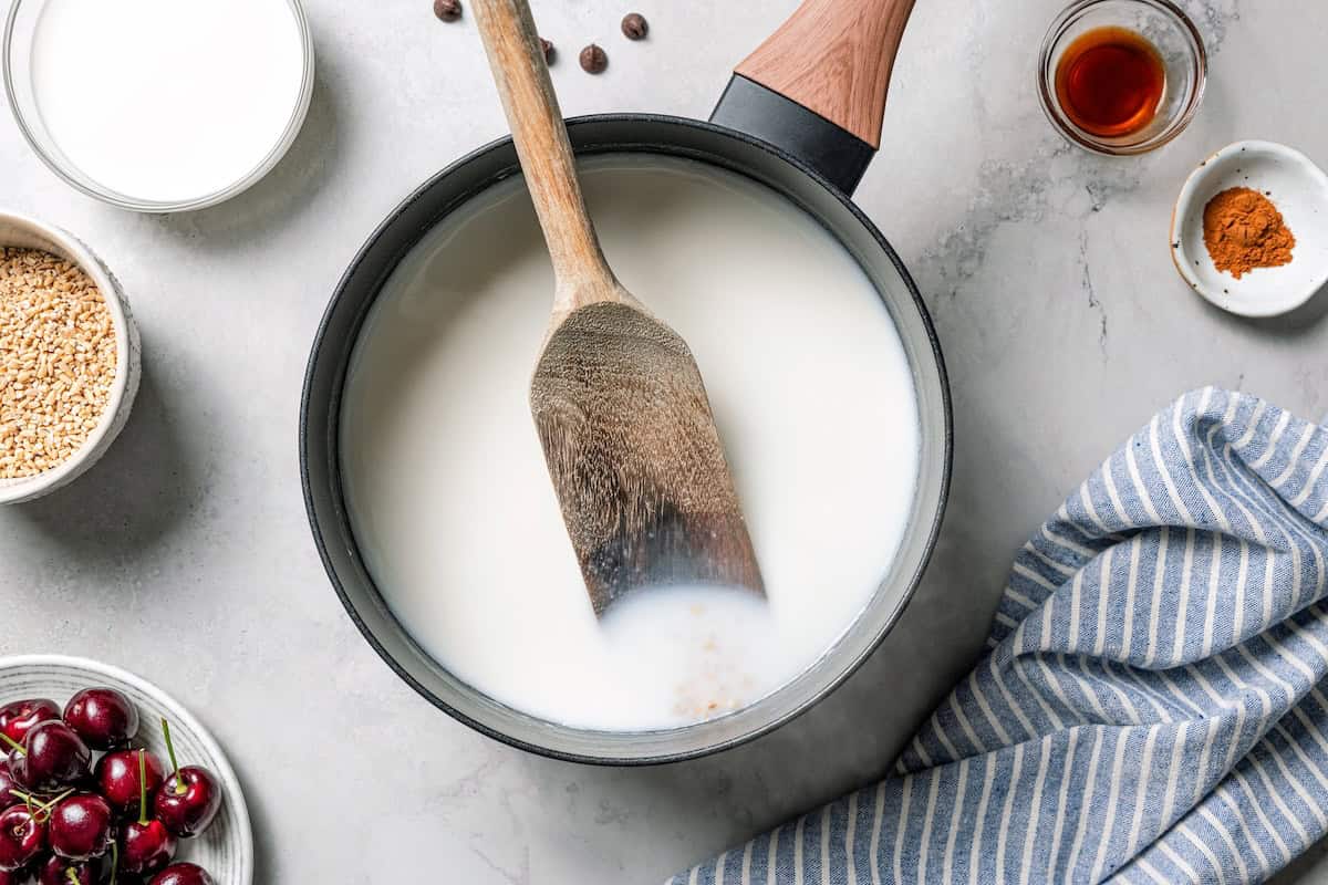 This easy overnight steel-cut oats recipe combines chewy oats, milk, vanilla, and cozy cinnamon into a delicious make-ahead breakfast. A spatula resting in a pot of steel cut oats cooking in milk.