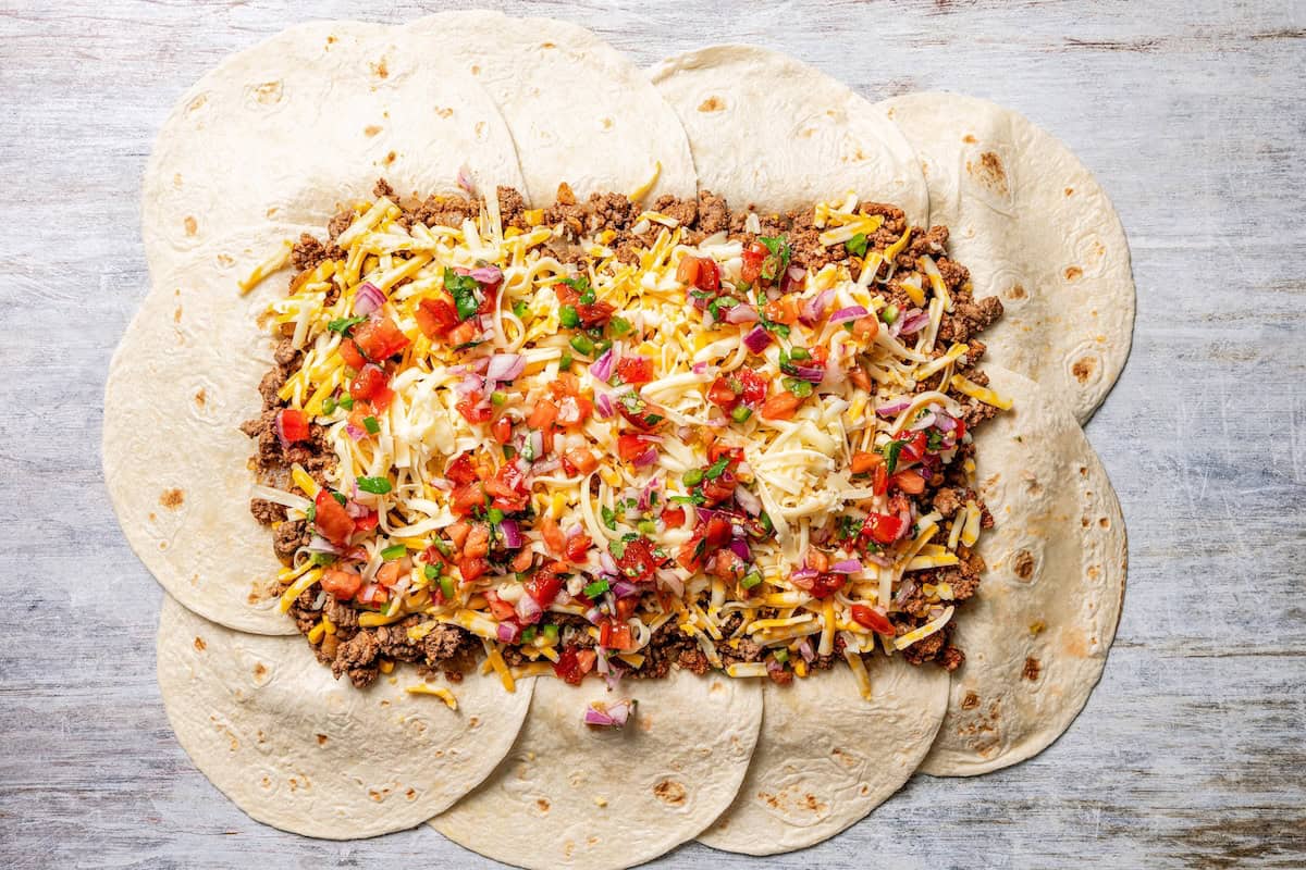 Baked sheet pan tacos are an easy way to make tacos for a crowd with crispy tortillas, seasoned ground beef, melty cheese, and pico de gallo. Taco ingredients layered on top of flour tortillas on a sheet pan.
