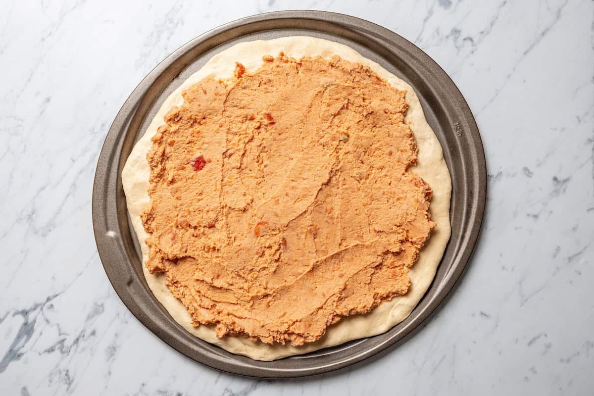 This Mexican-inspired taco pizza recipe has a crispy pizza crust topped with refried beans, taco meat, salsa, and your favorite taco toppings. Refried beans spread over a pizza crust.