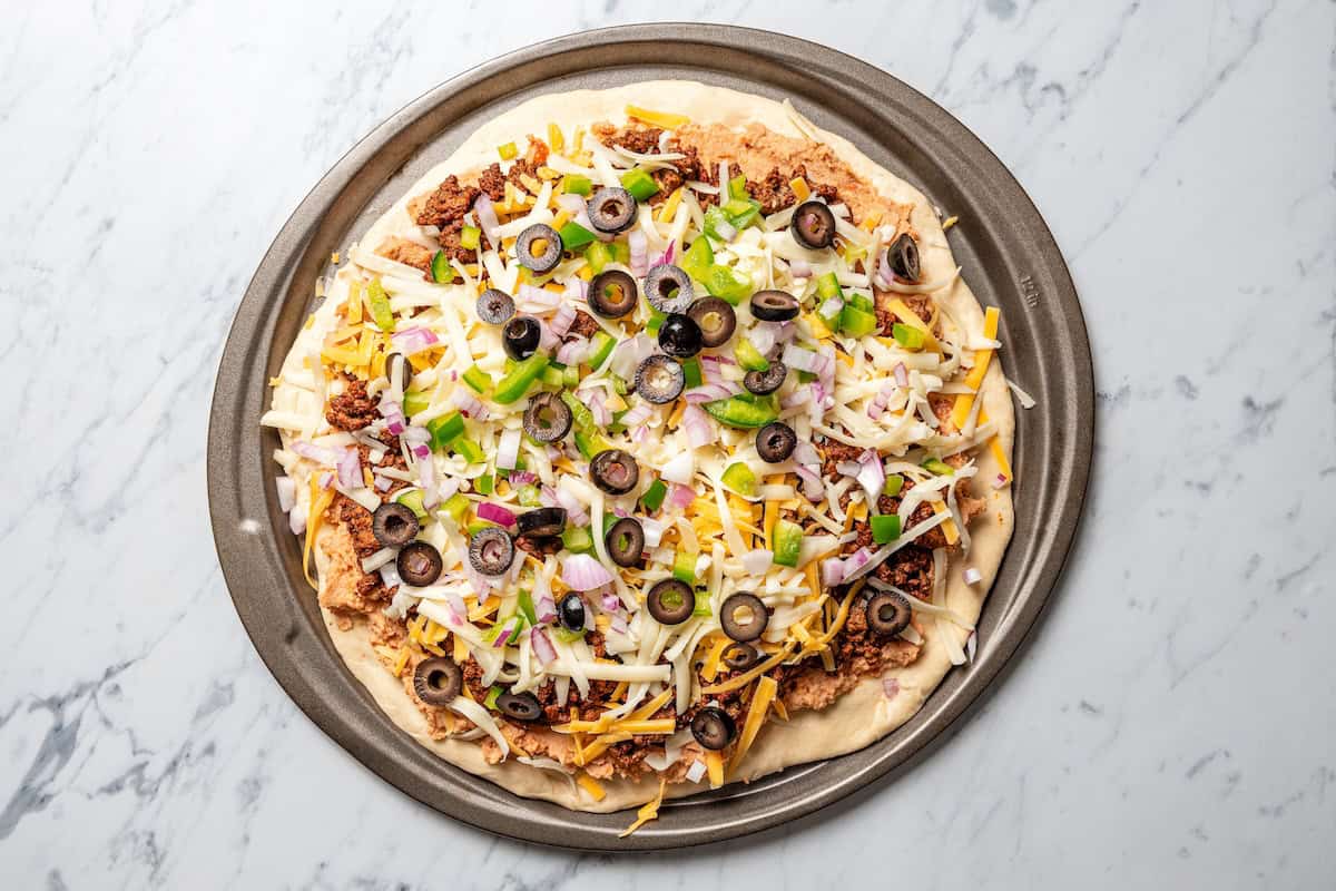 This Mexican-inspired taco pizza recipe has a crispy pizza crust topped with refried beans, taco meat, salsa, and your favorite taco toppings. Taco pizza ready to bake on a round pan.