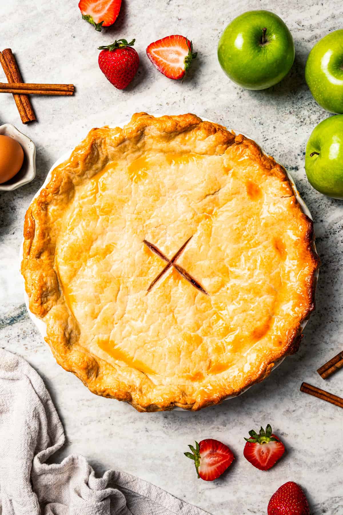 Apple berry pie is the perfect dessert to go from summer to fall! A cozy homemade apple pie bursting with fresh berries in a flaky pie crust. Golden-baked apple berry pie set on a table, with more fresh berries and apples arranged around the pie.