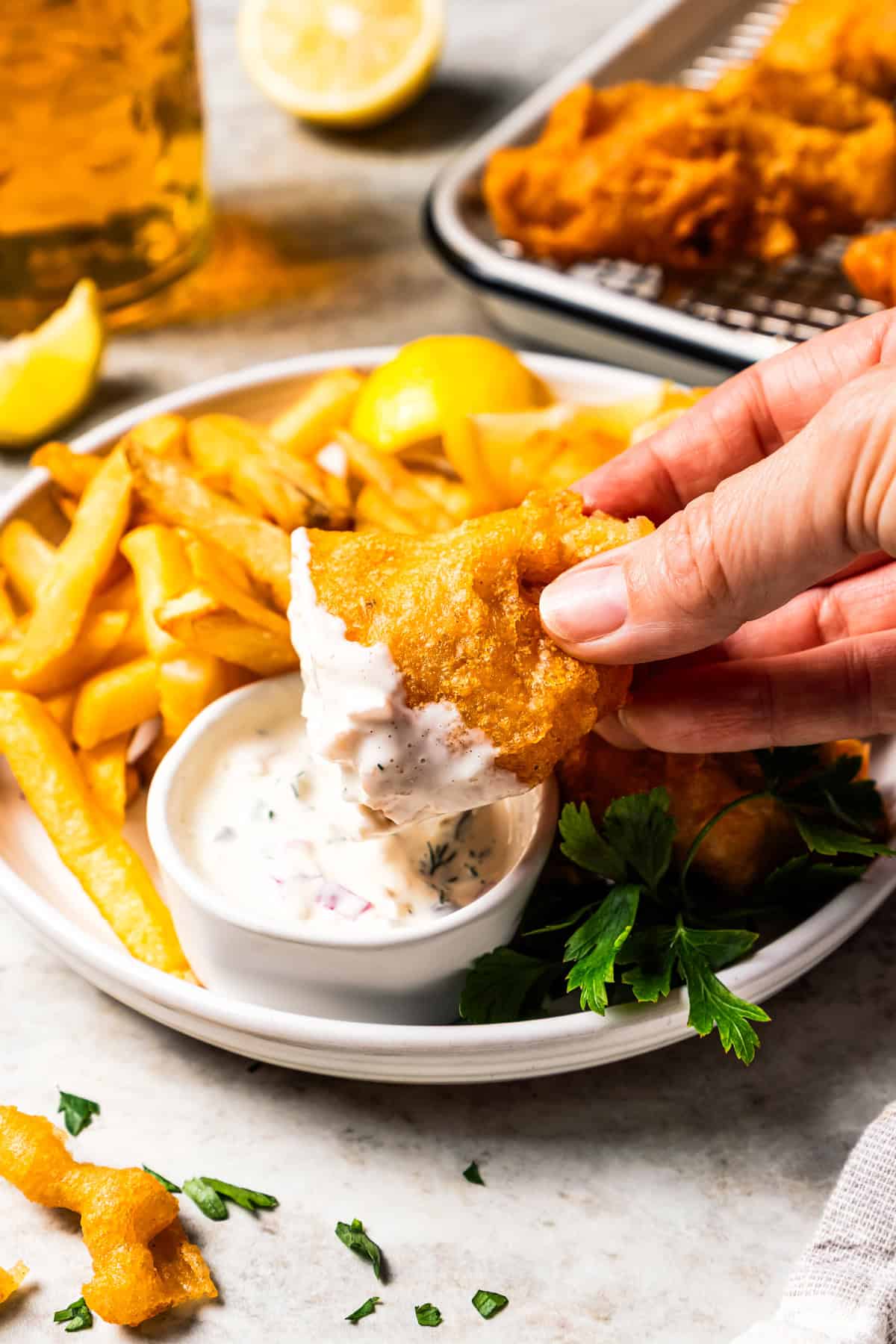 Make beer-battered fish with easy ingredients! This crispy, flaky fried white fish is perfect for tacos, pub-style fish and chips, and more. A hand dipping a piece of crispy beer battered fish into tartar sauce on a plate.