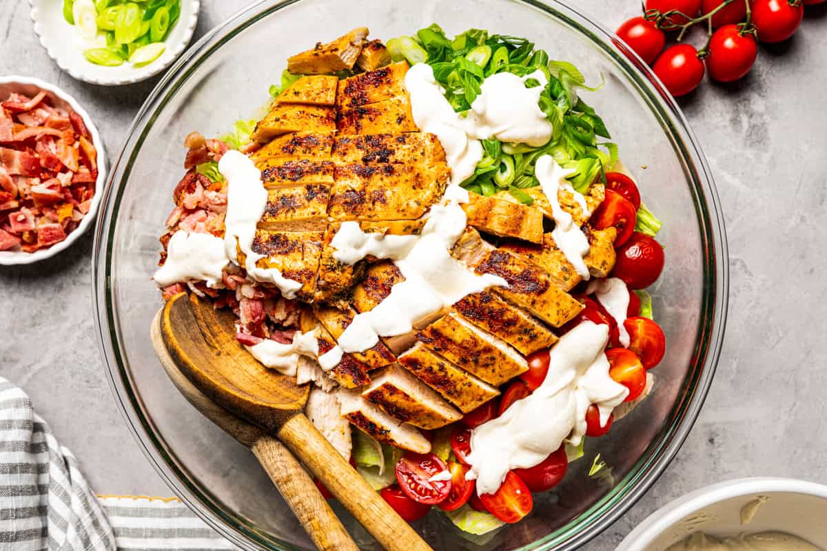 This easy BLT chicken salad combines juicy grilled chicken, crispy bacon, and ripe tomatoes with fresh lettuce and creamy yogurt dressing. Creamy salad dressing drizzled over BLT chicken salad ingredients in a bowl.