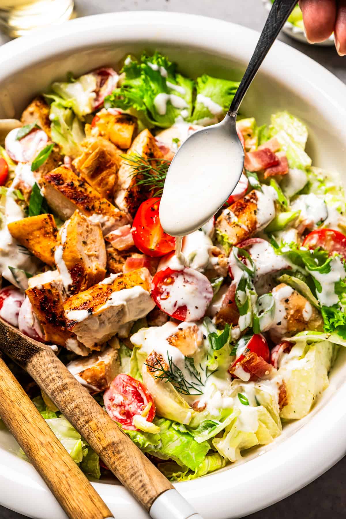 This easy BLT chicken salad combines juicy grilled chicken, crispy bacon, and ripe tomatoes with fresh lettuce and creamy yogurt dressing. Using a spoon to drizzle creamy dressing over a BLT salad with grilled chicken.